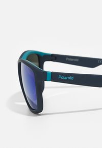 Occhiali da sole polarizzati con una montatura opaca di blu scuro, dettagli color verde acqua sulle asticelle e lenti viola sfumate. Presentano un marchio inciso.