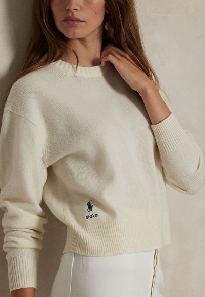 Donna che indossa un maglione di lana color crema con maniche lunghe e un piccolo logo Polo blu nella parte inferiore.