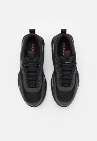 Sneaker neri con tomaia in materiali misti, caratterizzati da accenti in rete e sintetici, suola spessa e sistema di allacciatura prominente. Logo 'HUGO' visibile.