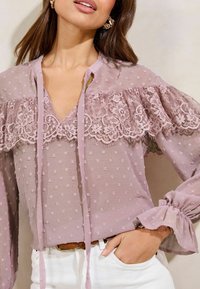 Light purple blouse featuring lace accents, dot texture, a V-neckline, and long sleeves with gathered cuffs. Ties at the neck complete the design.
