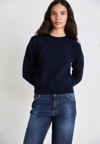 Suéter de punto azul marino con escote redondo y mangas acanaladas, combinado con jeans oscuros de piernas rectas. Diseño simple y limpio.
