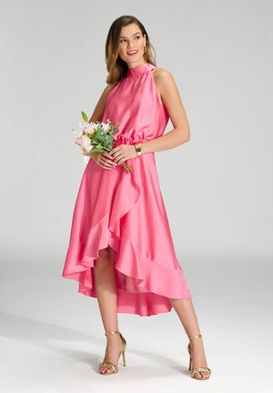 Pink satin dress with a halter neck, ruffled hem, and high-low design. Accented with gold heeled sandals and holding a bouquet.