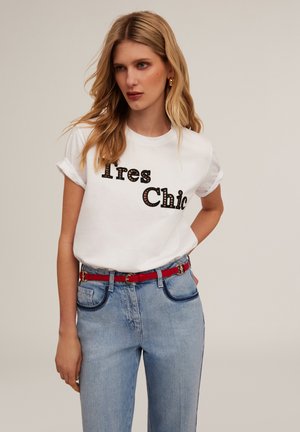 White cotton T-shirt with rolled sleeves featuring the text "Très Chic" in dark brown with a patterned outline; paired with light blue denim jeans and a red belt.