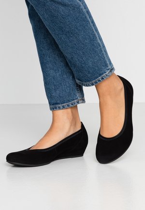 Piernas en jeans azules llevan bailarinas de tacón cuña de ante negro sobre un suelo suave de color gris claro contra un fondo liso.