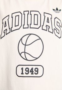 Fehér póló, amelyen hangsúlyos fekete "ADIDAS" felirat, egy kosárlabda grafika és alatta a "1949" felirat található. A sarokban fekete körvonalú trefoil logó is szerepel.