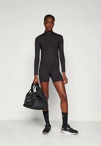 Idrettskvinne i svart langermet topp og shorts holder en svart treningsbag, iført svarte sokker og svart-rosa joggesko, stående mot en ensfarget bakgrunn.