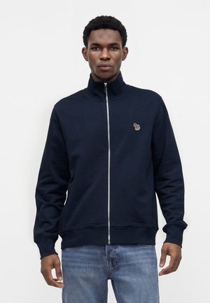 Jeune homme portant une veste zippée bleu marine avec un petit patch animal brodé sur la poitrine gauche, associée à un jean bleu.