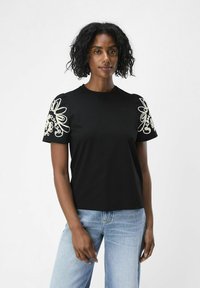 Svart t-shirt med intrikata vita blommönster broderade på korta ärmar, tillverkad av mjukt tyg; kombinerad med ljusblå jeans.