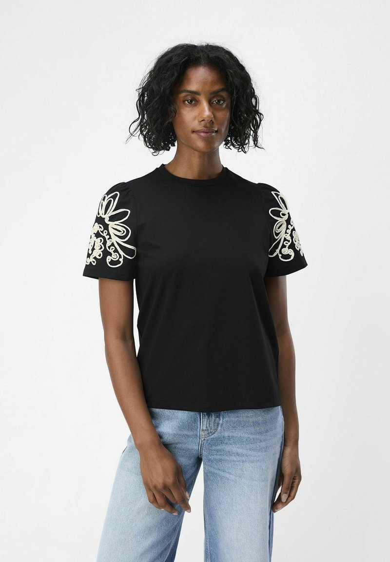 Svart t-shirt med intrikata vita blommönster broderade på korta ärmar, tillverkad av mjukt tyg; kombinerad med ljusblå jeans.