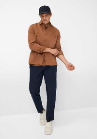Brun bomuldshemde med knaplukning, to forlommer og opsmøget ærmer, parret med marinebukser og hvide sneakers. Casual kasket på hovedet.