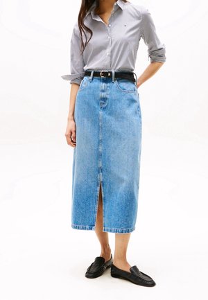 Kobieta ubrana w koszulę w paski zapinaną na guziki, midi spódnicę z denimu z wysokim stanem i przednim rozcięciem, czarny pasek oraz czarne wsuwane loafersy.