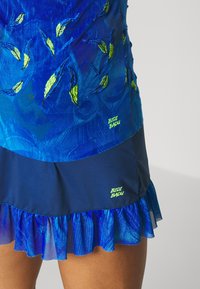 Modrá sukně s vyšívanými zelenými a modrými listy, volánkovým lemem a neonově zelenými logy "BIDI BADU" na tkaninových panelech.