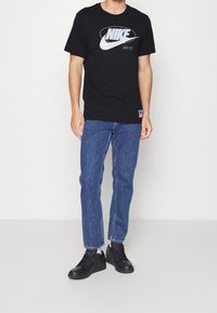 Schwarzes Baumwoll-T-Shirt mit dem "NIKE"-Logo in Weiß und Grün, kombiniert mit blauen Jeans und schwarzen Sneakers. Einfaches, lässiges Design.