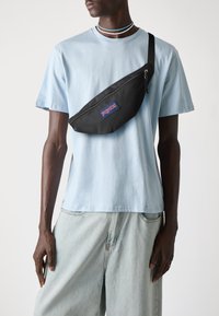 Borsetta in nylon nera con design curvo e tasca frontale con zip, caratterizzata da un logo "JanSport" blu e rosso, indossata sopra una maglietta azzurro chiaro.