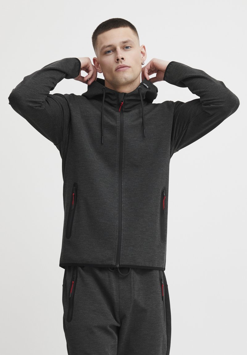 Veste à capuche zippée en tissu gris foncé avec une finition texturée, présentant des accents de zip rouges et des poches latérales, assortie à un pantalon correspondant.
