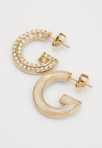 Naušnice u zlatnoj boji oblikovane poput slova "G", jedna s prozirnim rhinestones, a druga s glatkom, finom bojom sličnom slonovači.