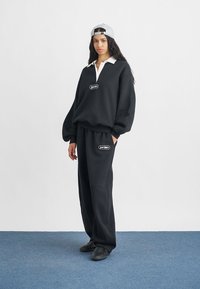 Μαύρη oversized φούτερ μπλούζα με λευκό γιακά και λογότυπο, συνδυασμένη με μαύρο παντελόνι φόρμας και ανοιχτό γκρι καπέλο. Κατάλληλη για καθημερινές εμφανίσεις.