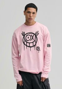 Hosszú ujjú rózsaszín pulcsi, amelyen fekete, graffitis stílusú grafika található az elülső részén. Az anyag lágy tapintású; a ujján logó folt található.