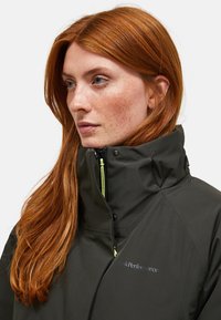 Eine rothaarige Frau mit Sommersprossen trägt eine dunkelgrüne Jacke mit hohem Kragen und Reißverschluss sowie einem "Peak Performance"-Logo auf der Vorderseite.