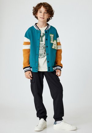 Teal en oranje varsityjack met gestreepte manchetten, met een "LA" patch en een grafisch t-shirt eronder. Zwarte cargobroek en witte sneakers.