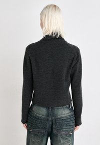 Pullover cinza de malha com gola alta e design cropped. Usado com calças de ganga azul com pregas texturizadas e detalhes em zíper.