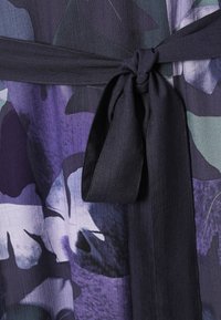 Közeli kép egy sötét lila szövetöv kötéséről, amelyet csomóba kötöttek lila, fekete és zöld virágmintás ruhadarab fölött.