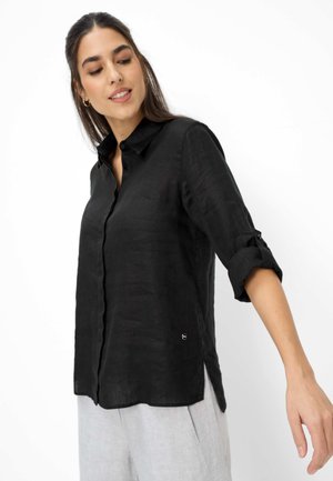 Frau trägt ein schwarzes Hemd mit aufgekrempelten Ärmeln und hellgrauen Hosen, schaut nach unten und streckt ihren rechten Arm aus.