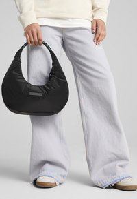 Sort nylontaske med en buet silhuet og et mønstret håndtag, som har en diskret logoaccent på fronten, holdt af en person.