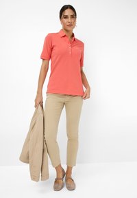 Polo shirt corallo con colletto e placket a tre bottoni, abbinato a pantaloni beige slim-fit e scarpe grigie basse. Tiene in mano una giacca color marrone chiaro.