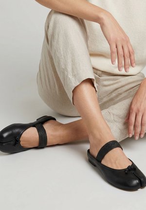 Person trägt beige Hose und cremefarbenes Oberteil, sitzt mit verschränkten Beinen und trägt schwarze geteilte Zehenleder Mary Jane Ballerinas mit gewebten Riemen.