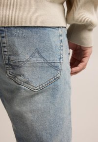 Light blue denim jeans with a textured finish and subtle distressed details, featuring a shaped back pocket with contrasting stitching.