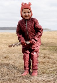 Vínová prošívaná bunda a kalhoty s šedými detaily, doplněné hnědým kloboukem s detaily na uších, držící peří venku.