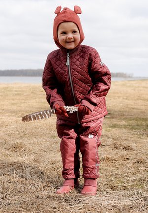 Bordeauxrote gesteppte Jacke und Overalls mit grauen Akzenten, kombiniert mit einem braunen Hut mit Ohrdetails, hält draußen eine Feder in der Hand.