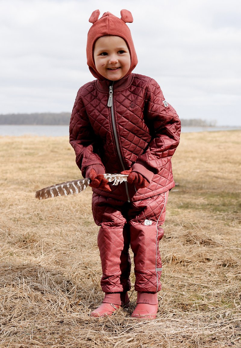 Vínová prošívaná bunda a kalhoty s šedými detaily, doplněné hnědým kloboukem s detaily na uších, držící peří venku.