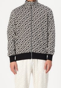 Chaqueta con cremallera negra y crema, con un patrón geométrico, puños y dobladillo acanalados, y un cuello alto. Mangas de largo completo, ajuste casual.