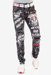 Schwarze Denim-Jeans mit distressed Details, Graffiti-ähnlichen Drucken, Totenkopf-Grafiken und roten Akzenten. Kombiniert mit einem weißen Gürtel und Sneakers.
