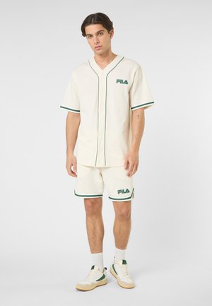 Man gekleed in een crèmekleurig FILA honkbalshirt en bijpassende short met groene afwerking, staand met een neutrale uitdrukking tegen een witte achtergrond.