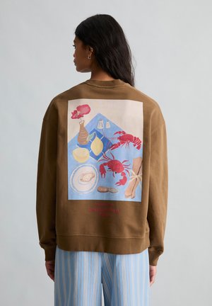 Kvinde iført brun sweatshirt med farverig illustration af flodkrebs og seafood på ryggen, kombineret med blåstribede bukser.