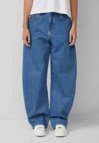 Plati zilu denim bikses ar augstu jostasvietu, kas aprīkotas ar klasisko piecu kabatu dizainu un baltām sporta apaviem. Gluda auduma tekstūra, ikdienišķs stils.