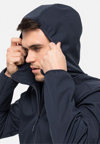 Sweat à zip bleu marine avec une coupe ajustée et une capuche réglable. Tissu lisse avec des détails de couture visibles, porté en ajustant la capuche.