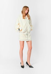 Sudadera de cremoso con cremallera y capucha grande, puños elásticos y falda corta a juego. Zapatos de tacón alto negros con adornos decorativos.