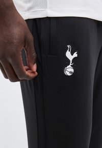 Gros plan d'une main à la peau foncée près d'un pantalon noir avec un logo d'oiseau blanc brodé posée sur un ballon de football.