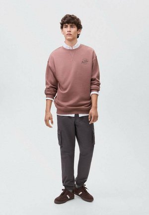 Felpa marrone con colletto bianco e logo, abbinata a pantaloni cargo scuri con tasche e polsini elasticizzati, indossata con sneakers marroni.