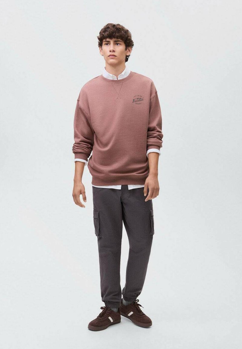 Felpa marrone con colletto bianco e logo, abbinata a pantaloni cargo scuri con tasche e polsini elasticizzati, indossata con sneakers marroni.