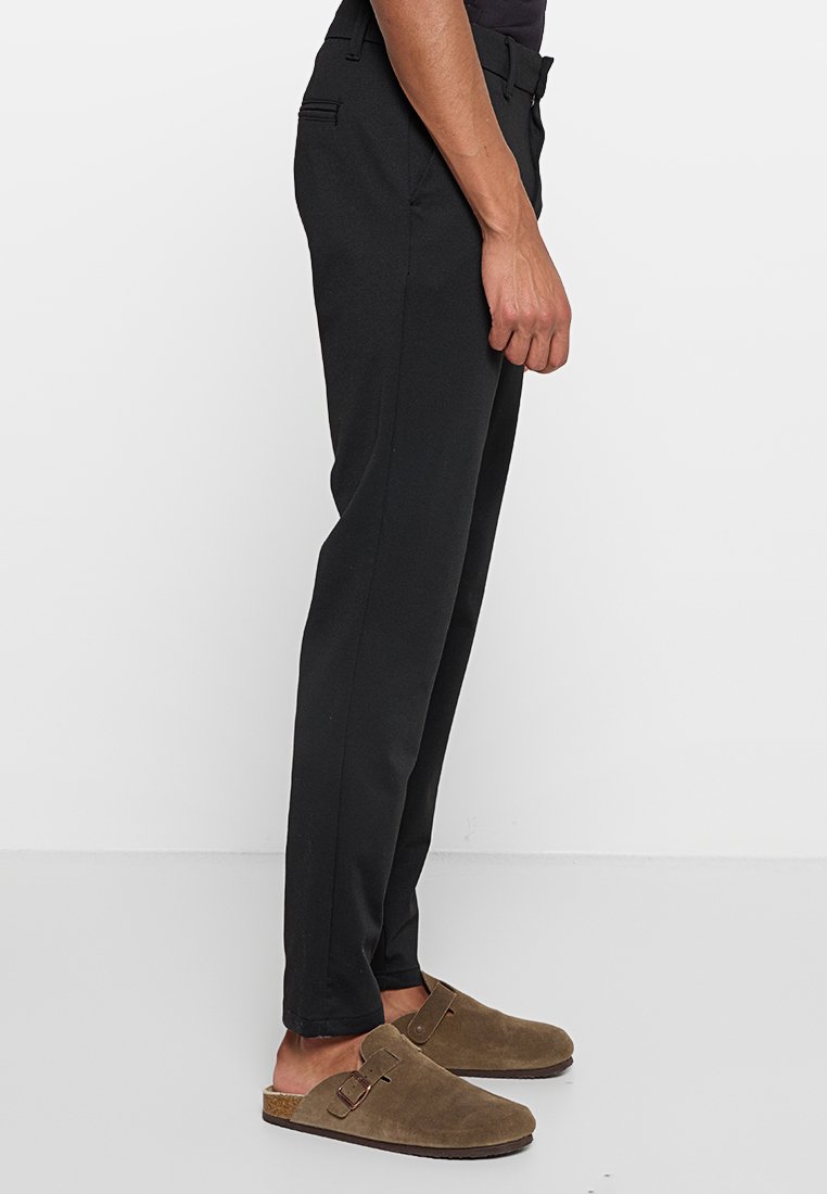Schwarze Hosen aus glattem Stoff, mit Slim-Fit, flachem Vorderteil, seitlichen Taschen und paspelierten Gesäßtaschen. Getragen mit braunen Slippers.