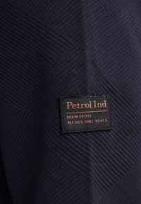 Tessuto blu navy con una texture a spina di pesce. Etichetta nera con la scritta "Petrol Ind" in arancione, con la dicitura "DENIM DRIVEN" e il numero di riferimento.