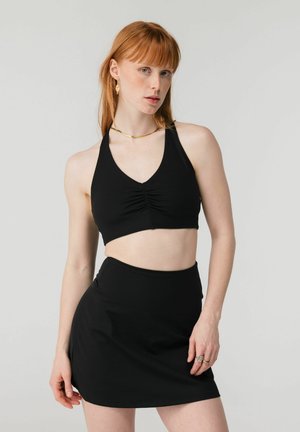 Rudovlasá žena v čiernom krátkom topu bez rukávov a čiernej sukni, stojaca pred jednoduchým svetlým pozadím.