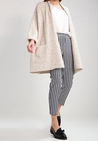 Beiger Cardigan mit lockerer Passform und seitlichen Taschen über einem weißen Hemd, kombiniert mit schwarz-weißen karierten Hosen und schwarzen Mokassins.