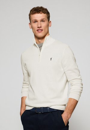 White, zip-neck sweater with ribbed cuffs and hem. Features a small black logo on the chest. Styled over a gray t-shirt, paired with navy pants.