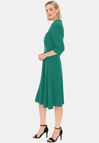 Turkusowa, midi sukienka z rękawami do łokcia, z metalowymi guzikami i rozkloszowanym dołem. Noszona z czarnymi, wysokimi sandałami na obcasie.